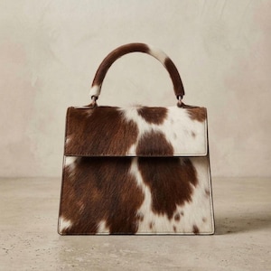 May include: A structured handbag featuring a cowhide exterior with a brown and white spotted pattern. The bag has a single top handle, a fold-over flap closure, and clean, straight edges, showcasing the natural texture of the animal hair.