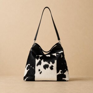May include: A hobo-style shoulder bag featuring a black and white cowhide pattern on the front panels. The bag includes a solid black leather top section, a matching black shoulder strap, and silver-toned metal hardware clips at the strap attachments.