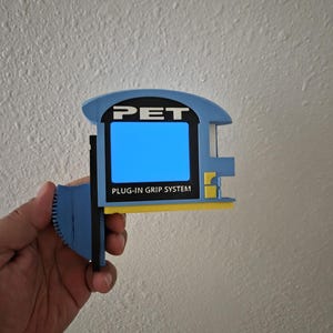 May include: A light blue, black, and yellow plastic device labeled PET PLUG-IN GRIP SYSTEM. The object features a rectangular blue center panel, a black vertical side component, and a textured blue circular grip area on the left side.