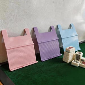 以下が含まれることがあります： パステルピンク、ラベンダー、ライトブルーのプラスチック製折りたたみ式ブックスタンド3点。それぞれリブ状の表面とヒンジ付きの支えが付いています。スタンドの横には、黒と赤の円形模様が描かれた白い麻雀牌が積み重なっています。