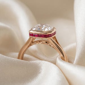 May include: A rose gold ring featuring a large cushion-cut clear centre stone surrounded by a halo of small, rectangular-cut red rubies. The band is crafted in rose gold with intricate, star-like engraved detailing along the upper shank.