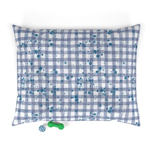 May include: A rectangular pet bed featuring a blue and white gingham check pattern overlaid with scattered dark blue paint-like splatters. A small blue spherical toy and a green dumbbell-shaped toy are positioned in front of the bed.