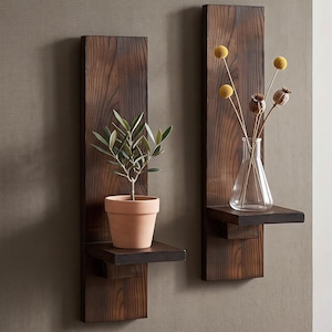 May include: Two vertical dark brown wooden wall-mounted shelves with rectangular backboards. One shelf holds a small green plant in a terracotta pot, while the other holds a clear glass vase containing yellow billy ball flowers and dried seed pods.