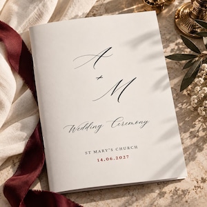 Può includere: Un libretto per cerimonia nuziale bianco con le iniziali calligrafiche A e M in nero, il testo Wedding Ceremony, St Mary's Church e la data 14.06.2027 stampata in rosso. Il libretto è presentato insieme a un nastro rosso scuro.