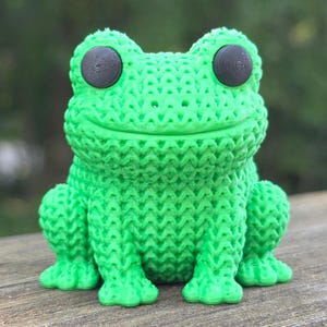 May include: A bright green frog figurine featuring a textured, woven-like surface pattern throughout its body. The frog has two large, circular, matte black eyes positioned on the top of its head and a wide, horizontal mouth line.