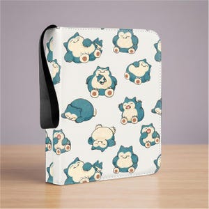 May include: A white zippered binder featuring a repeating pattern of blue and cream-colored Snorlax characters in various poses. The binder includes a black fabric wrist strap attached to the side, providing a secure way to carry the item.