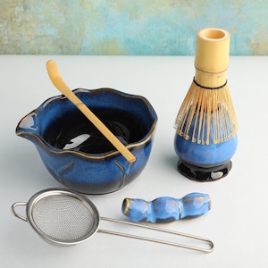 May include: A matcha tea set including a deep blue ceramic bowl with a scalloped rim, a bamboo whisk resting on a blue ceramic stand, a bamboo tea scoop, a metal mesh strainer, and a blue ceramic rest for the whisk or scoop on a white surface.