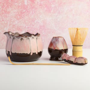 May include: A matcha tea set featuring a scalloped ceramic bowl with a pink and dark brown drip glaze, a matching whisk holder, a bamboo whisk, a bamboo tea scoop, and a small ceramic rest with a pink and dark brown drip glaze finish.
