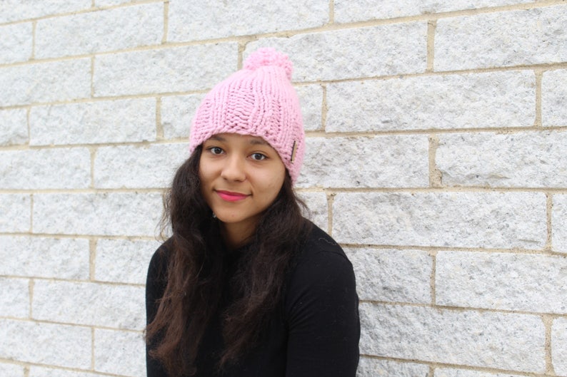 Puede incluir: Un gorro de punto rosa con un pomp&oacute;n esponjoso en la parte superior. El gorro lo lleva una persona con el pelo largo y oscuro.