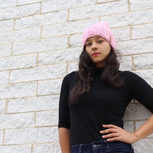 Puede incluir: Una mujer con una boina de punto rosa y una camisa negra de cuello alto con mangas largas. Est&aacute; de pie frente a una pared de ladrillo blanco y tiene la mano en la cadera.