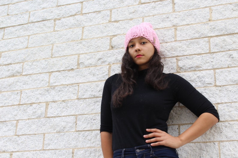 Puede incluir: Una mujer con una boina de punto rosa y una camisa negra de cuello alto con mangas largas. Est&aacute; de pie frente a una pared de ladrillo blanco y tiene la mano en la cadera.