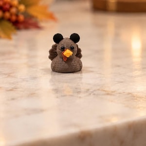 May include: A small, 3D-printed brown turkey figurine featuring two black circular ears on top of its head, a bright yellow beak, and a red wattle. The turkey has a textured, layered surface finish and two small black eyes.