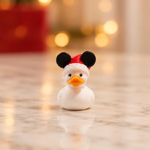 May include: A small white rubber duck figurine featuring an orange beak and black eyes. The duck wears a red and white Santa hat adorned with two black circular ears on top, creating a festive appearance with a smooth, matte-textured finish.