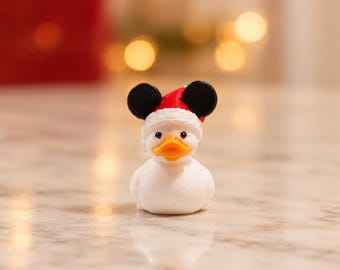 Puede incluir: Una pequeña figura de pato de goma blanco con pico naranja y ojos negros. El pato lleva un gorro de Papá Noel rojo y blanco adornado con dos orejas circulares negras en la parte superior, creando un aspecto festivo con acabado mate.