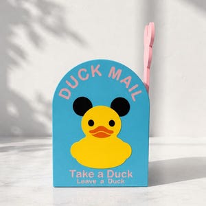 Può includere: Una cassetta delle lettere ad arco azzurra con un'immagine di un'anatra gialla con orecchie nere. Il testo DUCK MAIL è stampato in rosa in alto, e il testo Take a Duck Leave a Duck è stampato in rosa in basso. Una bandierina rosa sporge di lato.