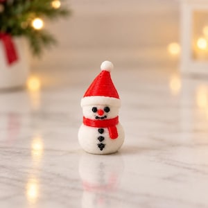 May include: A small white snowman figurine with a textured surface. The snowman wears a bright red hat with a white pom-pom and a matching red scarf. It features a small orange nose, black eyes, a black mouth, and three black buttons down its front.