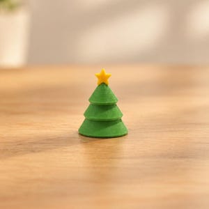 May include: A small, three-tiered green Christmas tree figurine with a smooth, layered texture. The tree is topped with a bright yellow five-pointed star. The object has a conical shape and a flat base, displaying a distinct 3D printed finish.