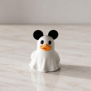 Puede incluir: Una pequeña figura blanca con forma de pato que lleva un disfraz de fantasma con un cuerpo de sábana blanca drapeada, dos orejas negras redondas en la parte superior, ojos negros circulares y un pico naranja texturizado.
