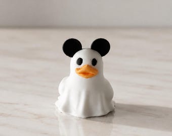 Könnte beinhalten: Eine kleine weiße Figur in Form eines Geistes, der über einer Ente drapiert ist, mit zwei runden schwarzen Ohren oben, schwarzen kreisförmigen Augen und einem orangefarbenen Schnabel. Das Objekt hat eine glatte, matte Oberfläche.