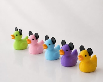 May include: A set of five small rubber ducks arranged in a row, each featuring a different body color including lime green, light pink, light blue, purple, and yellow. Each duck has a bright orange beak, black eyes, and two black rounded ears.