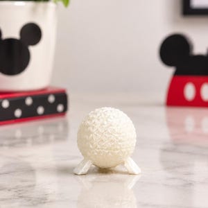 May include: A small, white, spherical object with a textured, geometric, triangular grid pattern on its surface. The sphere is supported by three short, angled legs, giving it a tripod-like appearance. The surface has a subtle, pearlescent sheen.