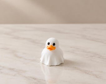 May include: A small, white, ghost-shaped figurine featuring the head of a duck with a bright orange beak and two round, black eyes. The figure is draped in a white sheet-like covering that flares out at the base, creating a smooth, rounded form.