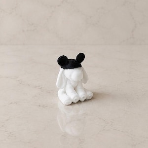May include: A small, white, 3D-printed figurine of an elephant sitting down. The elephant has large, floppy ears and is wearing a black, circular cap with two rounded black ears attached to the top, resembling a classic mouse-ear headpiece.