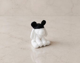 Puede incluir: Una pequeña figura blanca de un elefante en posición sentada. El elefante tiene grandes orejas caídas y lleva una gorra negra con dos orejas redondas negras unidas a la parte superior, que recuerda a un clásico gorro con orejas de ratón.