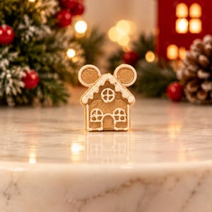May include: A light brown gingerbread house cookie with two circular ears at the top, decorated with white icing lines forming a roof, a central circular window, and two rectangular windows on the lower level, standing upright on a white surface.