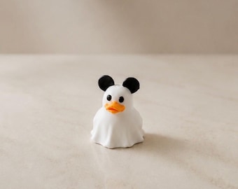 Könnte beinhalten: Eine kleine weiße Entenfigur, die ein weißes Laken über dem Körper trägt, um wie ein Geist auszusehen. Die Ente hat einen leuchtend orangefarbenen Schnabel, schwarze runde Augen und zwei abgerundete schwarze Ohren auf dem Kopf, die eine Maussilhouette nachahmen.