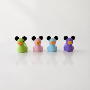Può includere: Quattro piccole statuette di paperelle di gomma disposte in fila. Ogni paperella presenta un colore del corpo pastello distinto in verde lime, rosa chiaro, azzurro e viola, abbinato a un becco arancione brillante, due piccoli occhi neri e due orecchie nere rotonde.
