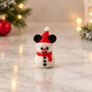 May include: A small white snowman figurine wearing a red Santa hat with black mouse ears, a red scarf, and three black buttons on its torso. The snowman has a small orange nose and black eyes, crafted with a visible layered texture.