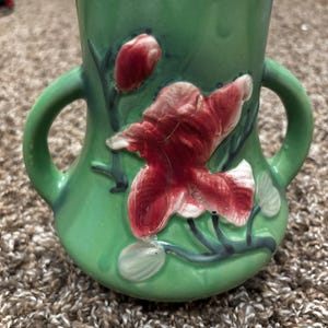 May include: A green ceramic vase featuring two side handles and a raised, three-dimensional relief of a large, blooming red flower with dark green stems and buds. The vase has a smooth, glossy finish and a wide, open top opening.