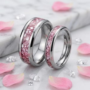 May include: Two silver-toned metal rings featuring a central inlay of crushed pink stone fragments. The rings have a polished, reflective finish and a smooth, rounded band design, showcasing a consistent aesthetic across both pieces.