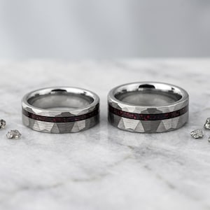 Puede incluir: Dos anillos de metal plateado con una textura exterior martillada y facetada. Cada anillo presenta una incrustación horizontal central de material triturado de color rojo oscuro y brillante que contrasta con el acabado metálico.