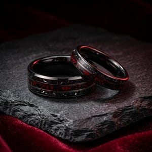 Puede incluir: Dos anillos negros con una incrustación central de material rojo y negro triturado. El anillo más ancho tiene una banda roja prominente flanqueada por bordes negros, mientras que el anillo más fino muestra un patrón moteado rojo y negro.