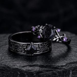 May include: A black metal ring set featuring a band with a square-cut dark blue sandstone and leaf engravings, paired with a ring showcasing an oval-cut dark blue sandstone centre stone accented by small purple round stones and leaf-shaped metal details.