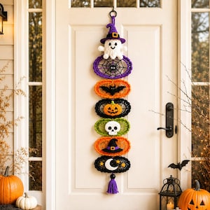May include: A vertical Halloween wall hanging featuring a series of six crocheted oval motifs connected in a stack. The design includes a white ghost in a purple hat, a black spider web, a black bat, an orange pumpkin, a white skull, a black witch hat, and a white crescent moon with stars, finished with a purple tassel.