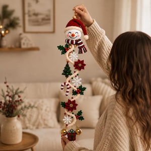 Op de afbeelding: Een verticale gehaakte kerstwandhanger met bovenaan een sneeuwpop met een rode muts en een gestreepte sjaal. De crèmekleurige spiraalvormige basis is versierd met gehaakte kerststerren, hulstblaadjes, een kleine groene boom en witte sneeuwvlokken.