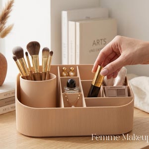 Pode incluir: Um organizador de maquilhagem bege com textura canelada, apresentando um suporte cilíndrico para pincéis e vários compartimentos retangulares. Contém pincéis de maquilhagem com ponteiras douradas, frascos de perfume, joias e sombras.