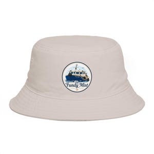 May include: A light beige bucket hat featuring a circular graphic on the front. The graphic depicts a blue and white fishing boat on water with the text The Fundy Mist written in a dark blue script font inside the circular border.