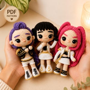 May include: Three amigurumi dolls with large black eyes and rosy cheeks. The left doll has purple hair, the centre doll has black hair, and the right doll has pink hair. They wear white and black outfits with gold accents. A PDF pattern label is visible.