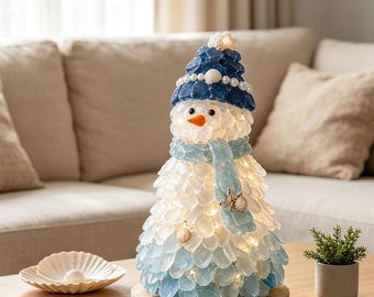 May include: A snowman figurine crafted from translucent white and light blue sea glass pieces, featuring a dark blue hat adorned with white pearls and a small shell, a light blue scarf with a starfish, and an orange nose, illuminated by internal lights.
