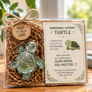 May include: A clear glass turtle figurine with the word MASON embossed on its shell, packaged in a clear box with brown crinkle paper, a twine bow, and a tag reading MADE with CARE. A card features a turtle illustration and text: EMOTIONAL SUPPORT TURTLE. I used to be a mason jar. Life changed. So I did. Now I'm here to remind you—SLOW DOWN, YOU MATTER. One day at a time, friend. You're doing better than you think.