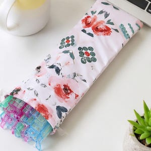 May include: A rectangular fabric pouch featuring a light pink background with a floral pattern of red and pink roses, green leaves, and small dark green geometric motifs. The pouch is partially open, revealing several translucent blue plastic clips.