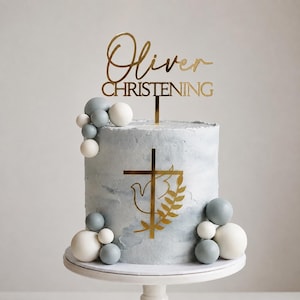 Puede incluir: Un pastel cilíndrico con glaseado azul claro texturizado, decorado con grupos de esferas blancas y grises. Un adorno dorado para pastel dice Oliver CHRISTENING, y un emblema de cruz dorada con una paloma y una rama está centrado.