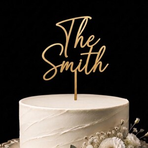 Könnte beinhalten: Ein goldfarbener Tortenstecker mit dem Schriftzug The Smith in Schreibschrift. Der Tortenstecker ist auf einem dünnen vertikalen Stab befestigt und steckt in einer weiß glasierten Torte, die mit kleinen weißen Blüten und grünen Blättern verziert ist.