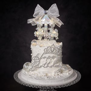 May include: A white cake decorated with white pearls and clear spheres, featuring a silver rhinestone Happy Birthday topper and a carousel-themed cake topper with clear horses, a central pillar, and a large sheer white organza bow on top.