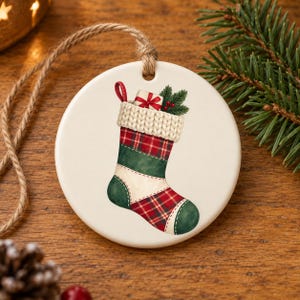May include: A round white ceramic ornament featuring a printed illustration of a Christmas stocking. The stocking has a cream-colored knit cuff, red and green plaid sections, and a solid green heel and toe. A small gift box and pine sprigs are tucked inside.