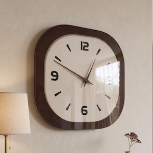 Puede incluir: Un reloj de pared cuadrado con esquinas redondeadas, que presenta un marco marrón oscuro y una esfera de color crema. El reloj tiene números negros en las posiciones 12, 3, 6 y 9, marcas negras para las otras horas y manecillas negras finas.